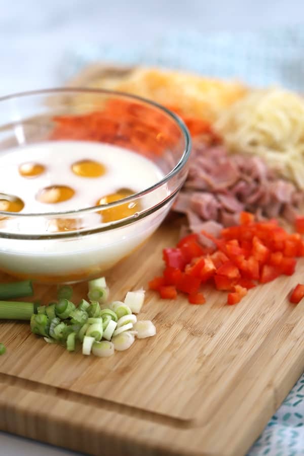 a glass bowl filled with milk and eggs, sitting on a cutting board with chopped green onions, red peppers, ham and cheese