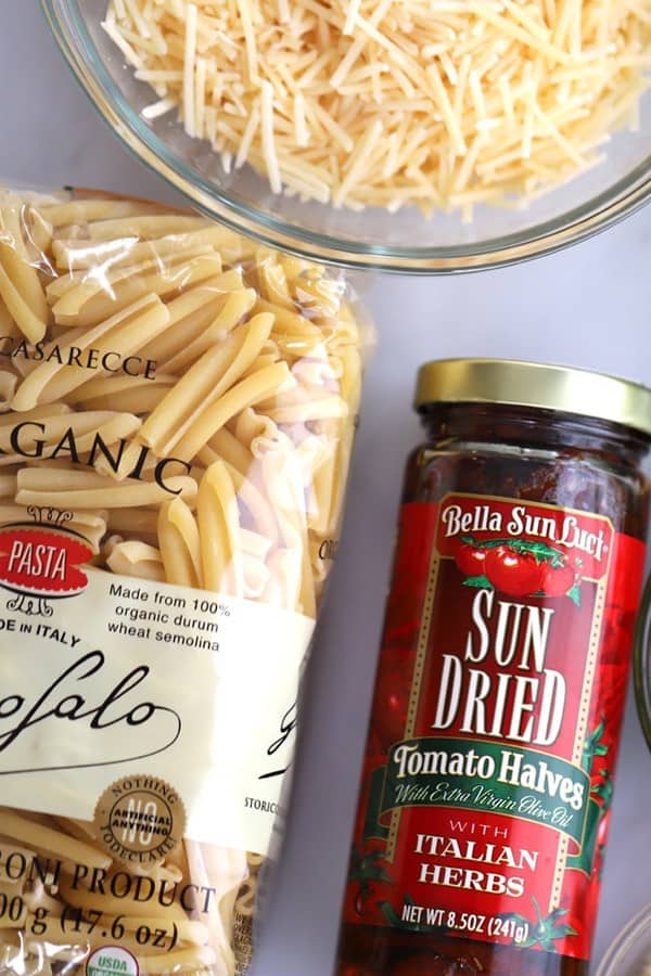 A bag of dried pasta, a jar of sun-dried tomatoes and a glass bowl full of shredded cheese on a tabletop. Chicken sun dried tomato pasta bake.
