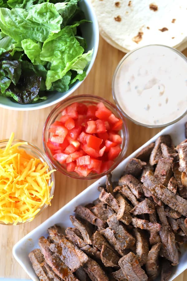 Carne asada chipotle flavored meat on a cutting board with cheese, tomatoes, lettuce and chipotle ranch, how to make burritos, asada tacos, tacos de asada, chipotle meat options. 