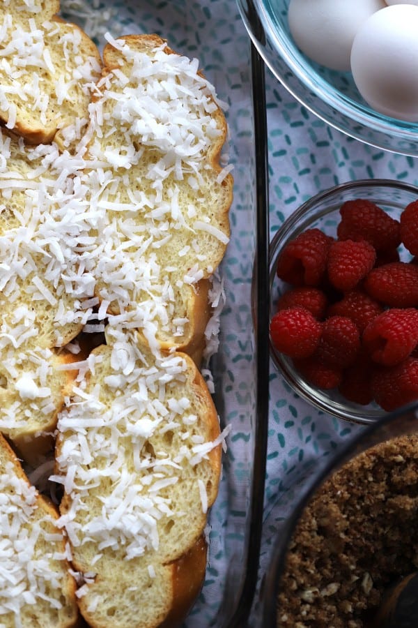 french bread slices topped with coconut flakes in a glass baking pan, with ramekins of raspberries and eggs beside