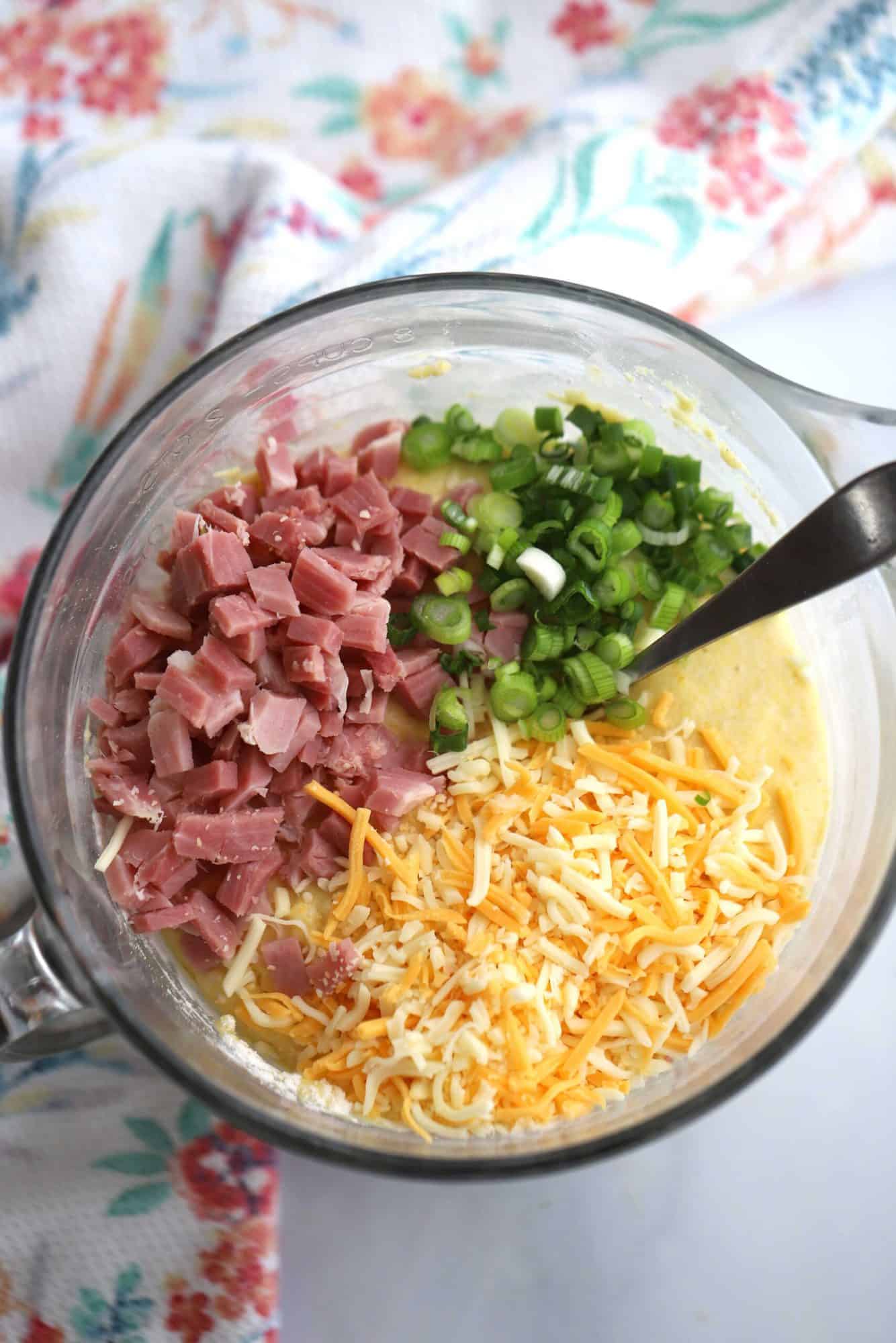 Ham and Cheese Corn Muffin batter in a pitcher with chopped ham, sliced onions and shredded cheese on top ready to mix in. cheese and ham muffins. 