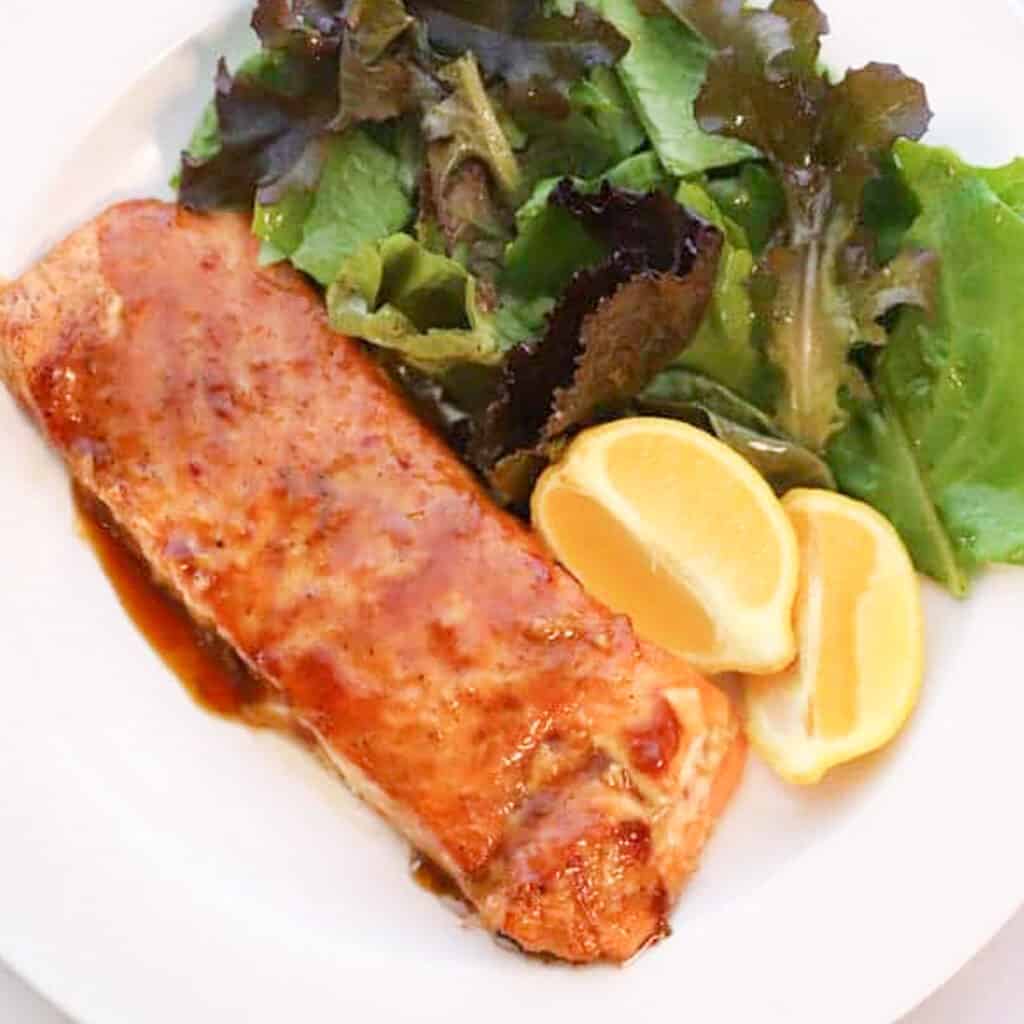 Sweet teriyaki Glazed Salmon on a white dinner plate with a side salad and lemon wedges.