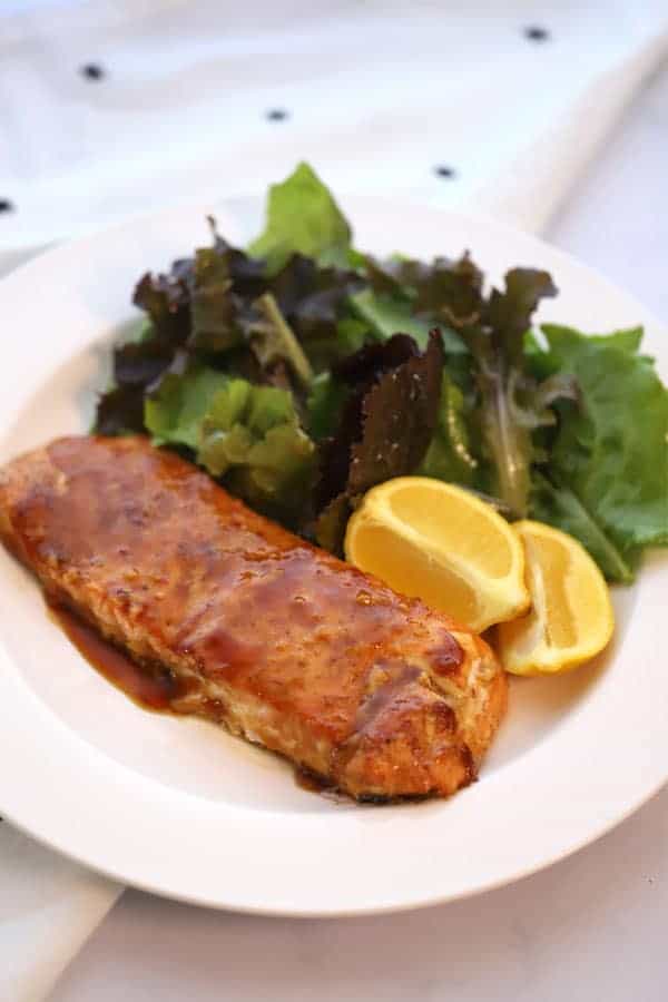 A white serving plate with a portion of glazed salmon and fresh lemon wedges with a side salad.