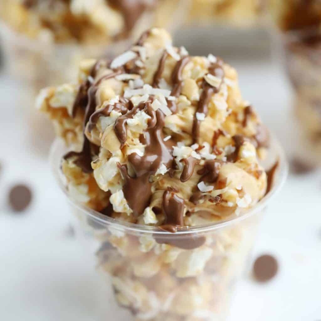 A plastic cup filled with caramel popcorn, topped with coconut and chocolate sauce.