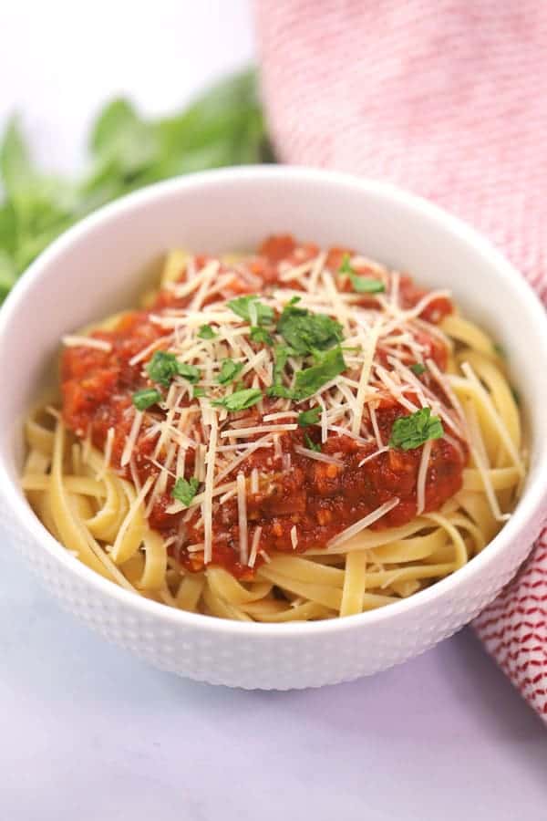 A white bowl full of fettuccini noodles topped with marinara sauce, Parmesan cheese and fresh basil.
