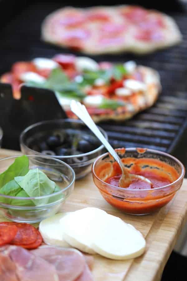A grill cooking Grilled Pizza and a cutting board to the side with pizza toppings. Pizza dough grill, grilled pizza recipes, pizza dough for grill.