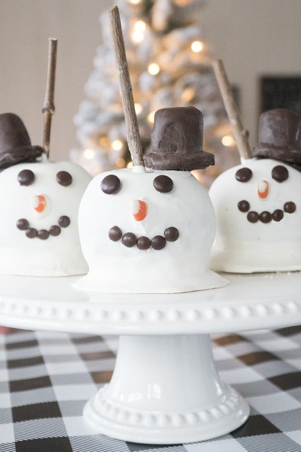 Three Snowman Christmas Candy Apples on a cake stand on a table with a checkered tablecloth. Christmas caramel apples, easy caramel recipe.