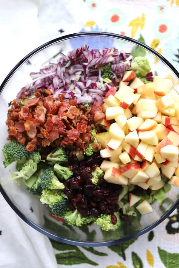 A glass bowl full of ingredients to make Broccoli Salad with Bacon raisins.