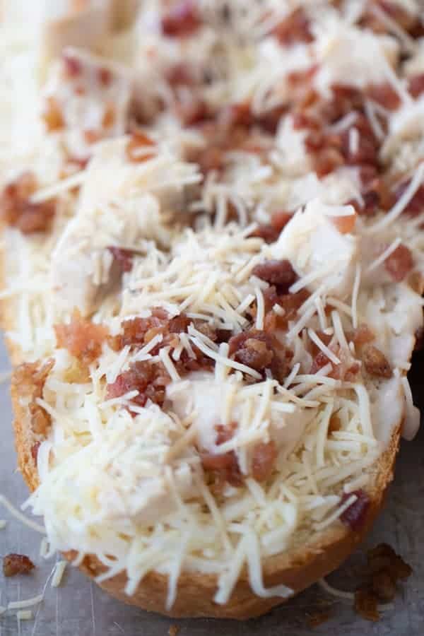 A loaf of french bread cut in half and topped with cheese, bacon and chicken, ready to be cooked in the oven.