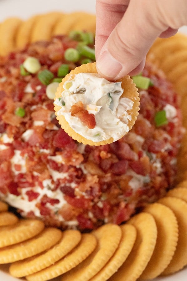 A hand holding a cracker dipped into Jalapeno Popper cheese ball, with the entire appetizer and crackers in the background; bacon cheese ball.
