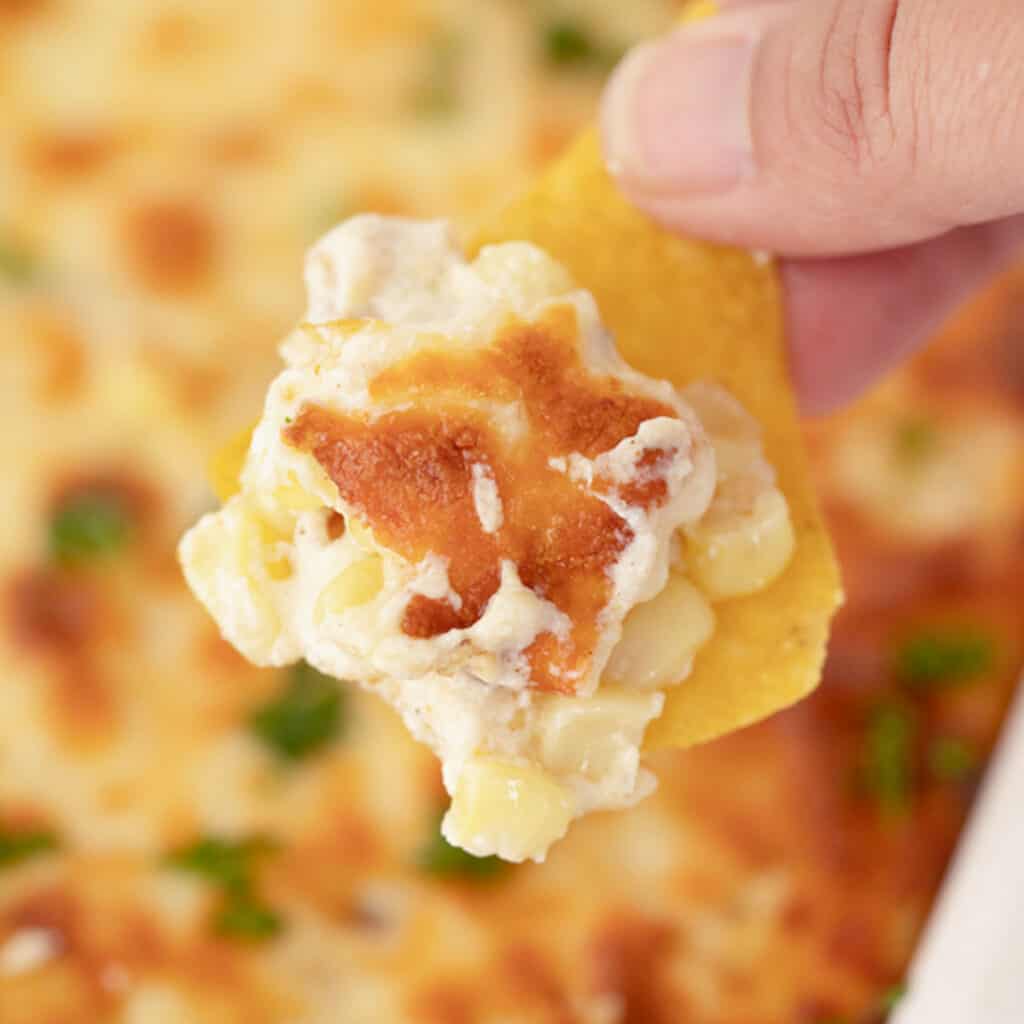 Easy Fiesta Corn Dip in a baking dish with a hand dipping a chip into it. Best easy appetizers.