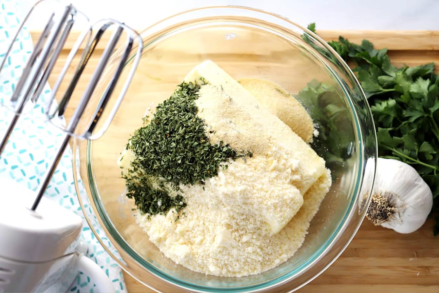 Butter, garlic powder, dried parsley and Parmesan cheese in a glass bowl with a hand mixer next to it.