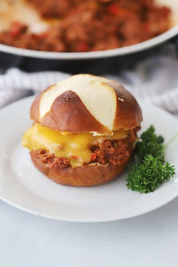 a white plate with sloppy joes on a pretzel bun with melted cheese and a sprig of parsley