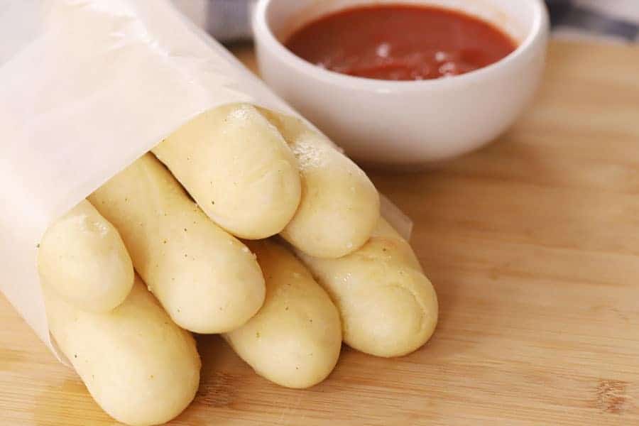 breadsticks olive garden wrapped in a sheet of parchment next to a small bowl of marinara dipping sauce, copycat olive garden breadsticks, best breadstick recipe.