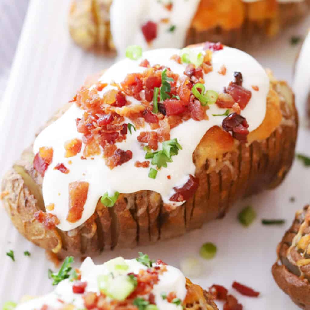 Loaded hasselback potatoes on a white plate covered in sour cream, bacon, and green onions.  Valentine dinner at home. 