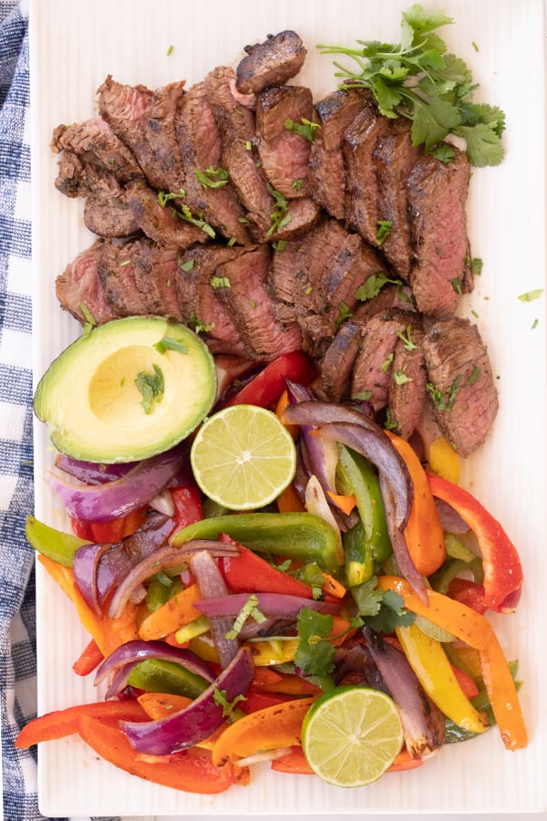 Steak fajitas sliced on a white plate along with veggies, avocado and lime.