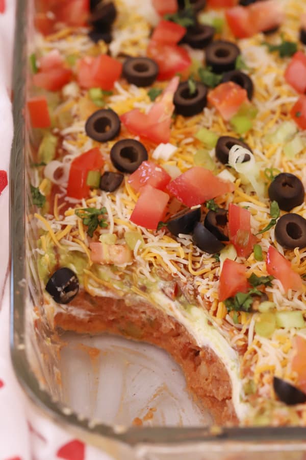 A layered bean dip recipe in a baking dish with a portion removed so you can see the layers, seven layer bean dip, bean dip layer. 
