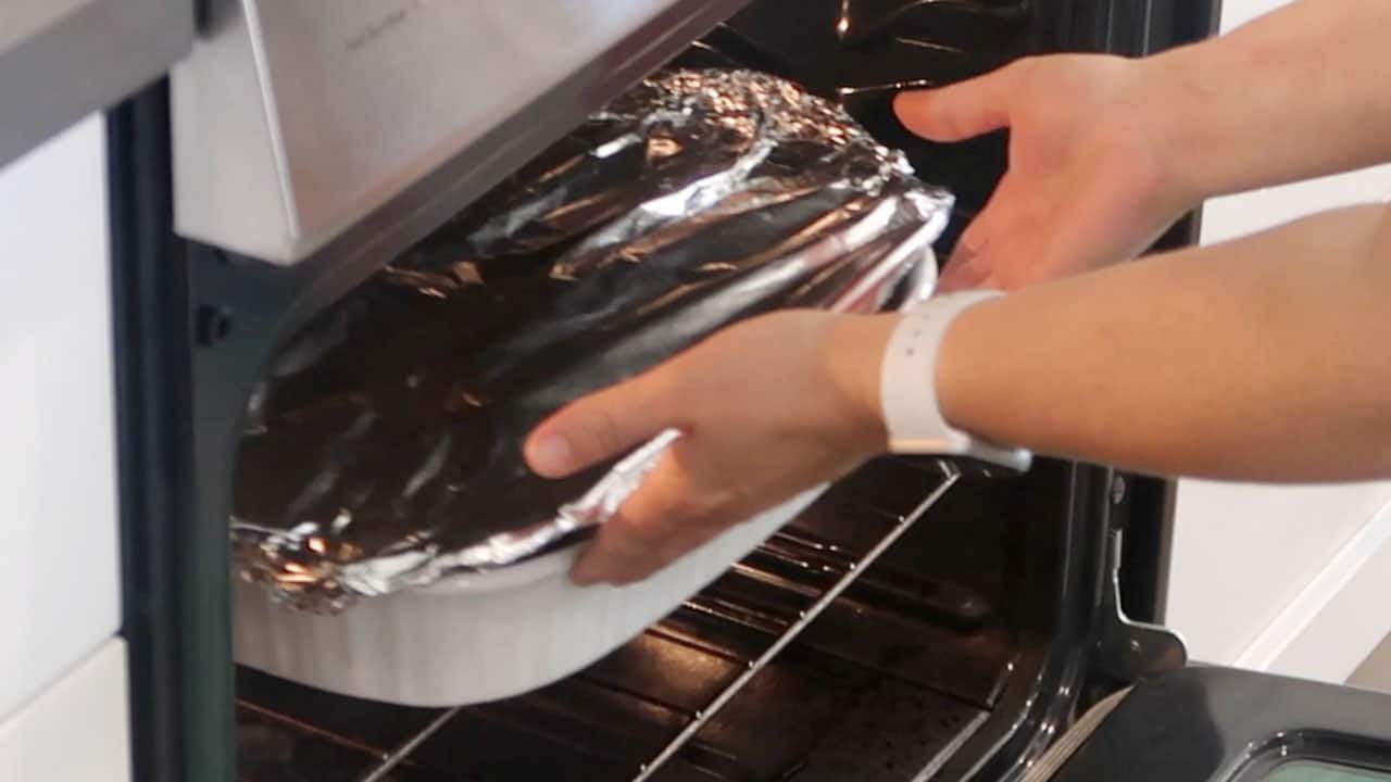 Hands placing a baking dish covered with aluminum foil into an oven.