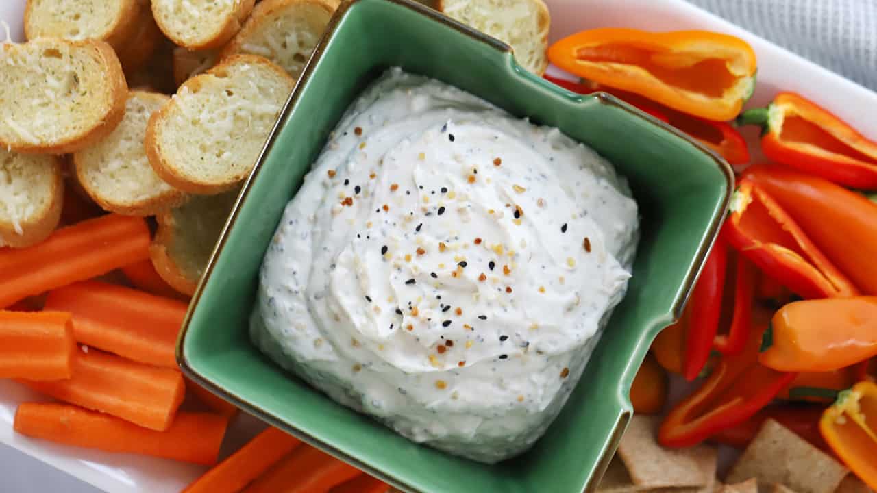 Everything bagel dip in a serving dish surrounded by crostini and fresh veggies.