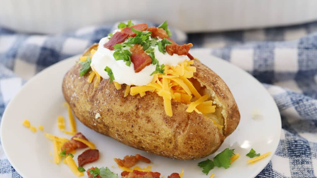 A white serving plate with a baked potato served restaurant style—fully loaded with sour cream, bacon, cheese, and more.