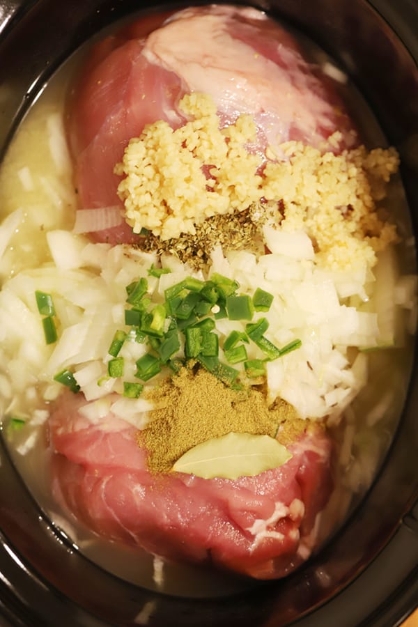 A slow cooker filled with a raw pork roast, broth, garlic, onions, jalapenos and seasoning.