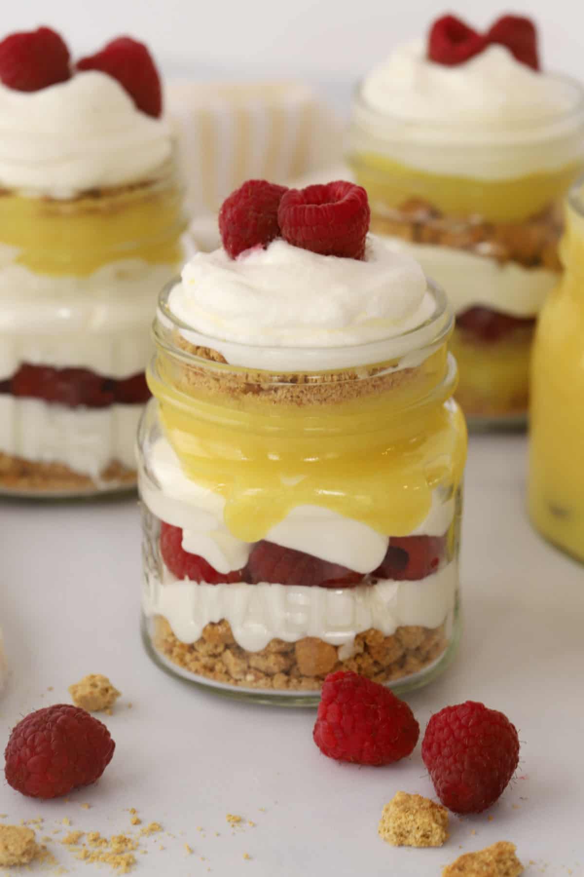 Parfaits built in small glass mason jars, with layers of curd, whipped cream, raspberries and cookies.