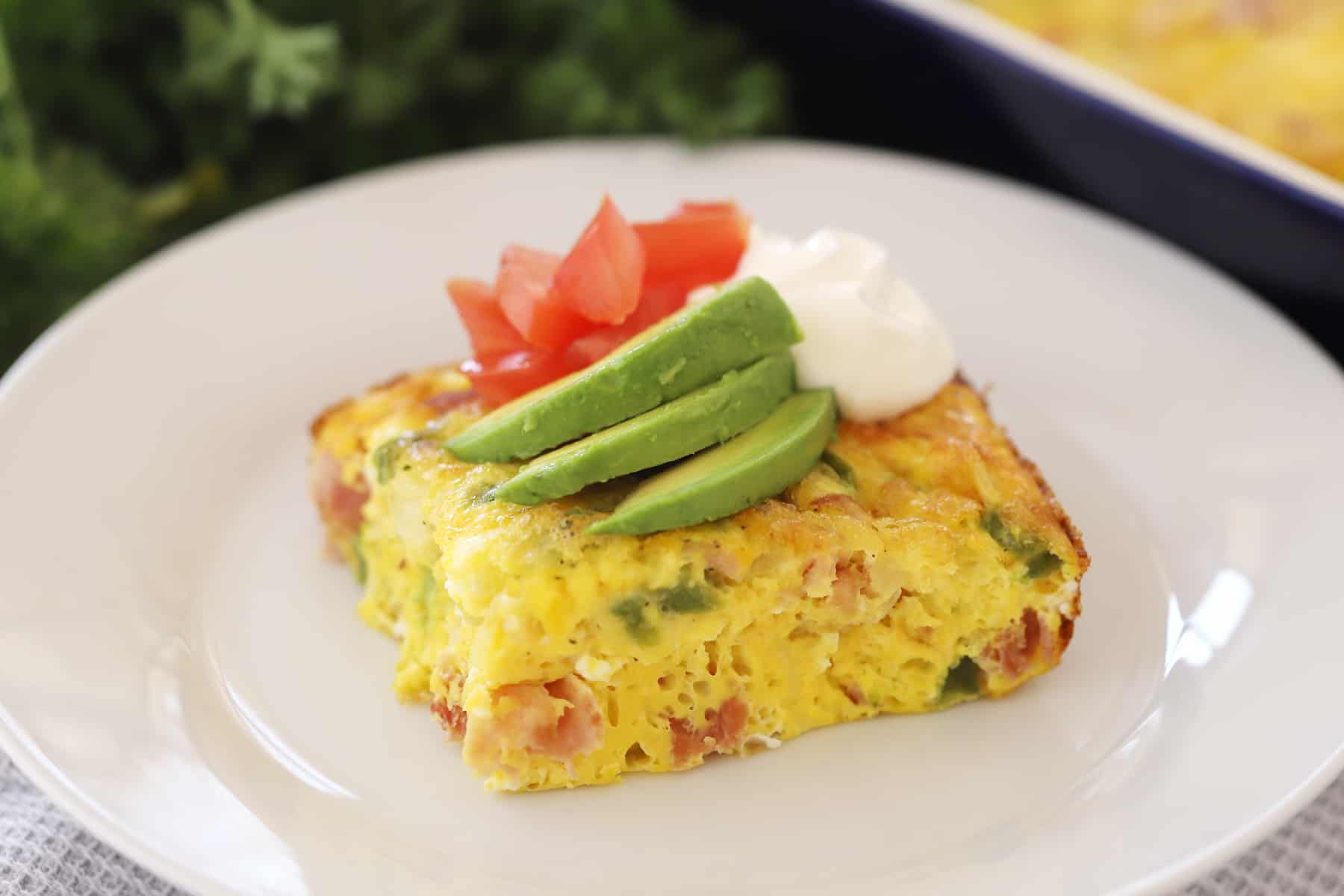 A slice of baked omelet on a white plate topped with sliced avocado, diced tomatoes and a dollop of sour cream.