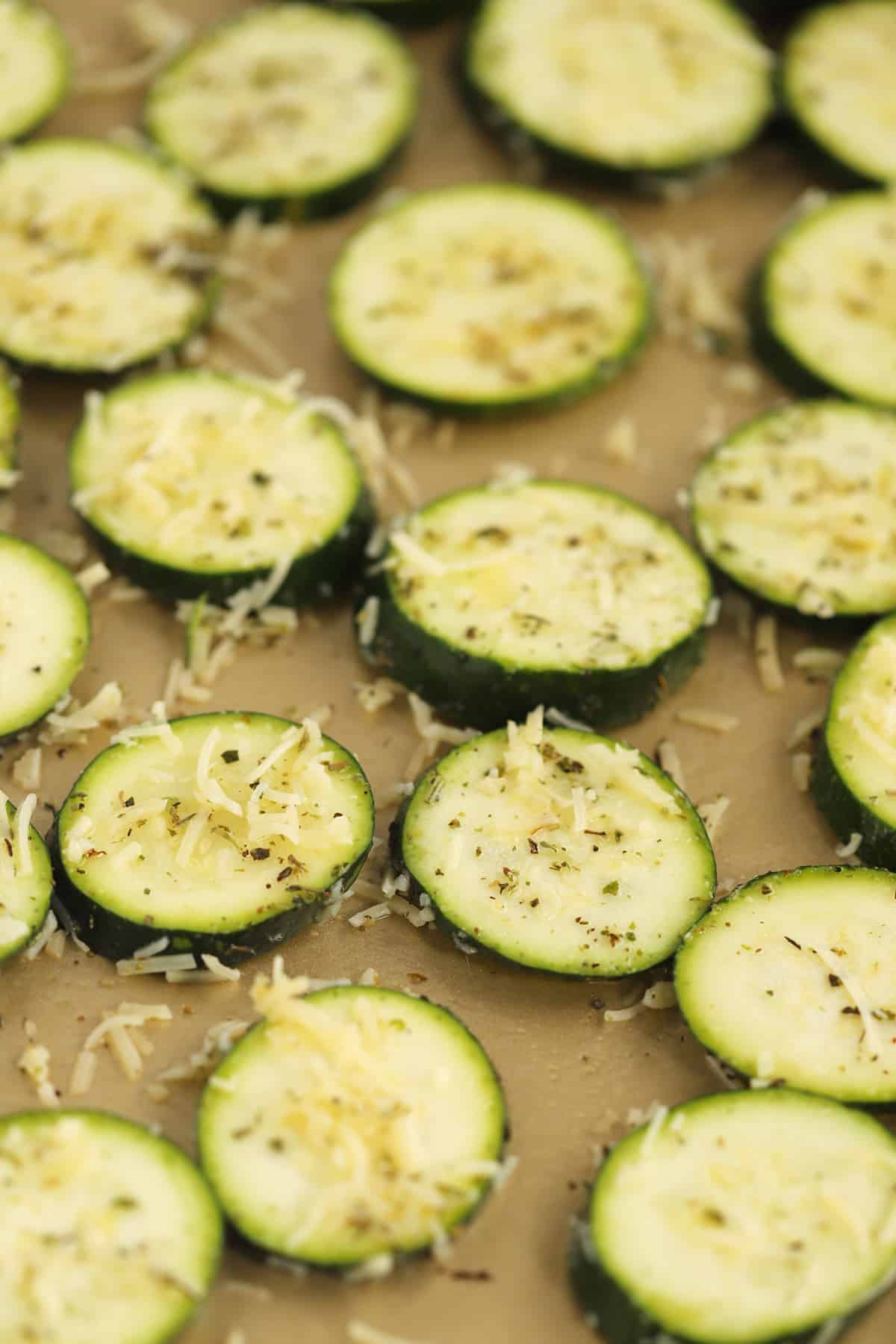 Thinly sliced rounds of zucchini on a sheet tray covered with seasoning and Parmesan cheese.
