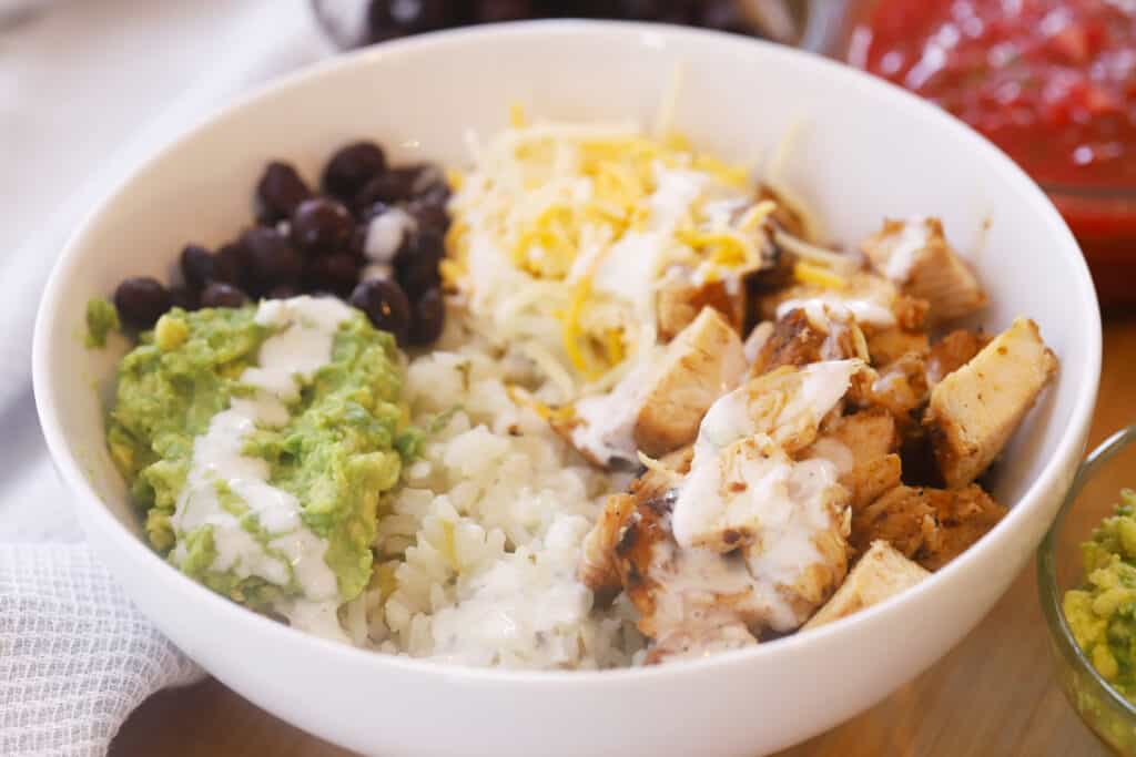 A white bowl filled with rice, chicken, beans, guacamole and shredded cheese. chipotle burrito bowl recipe.
