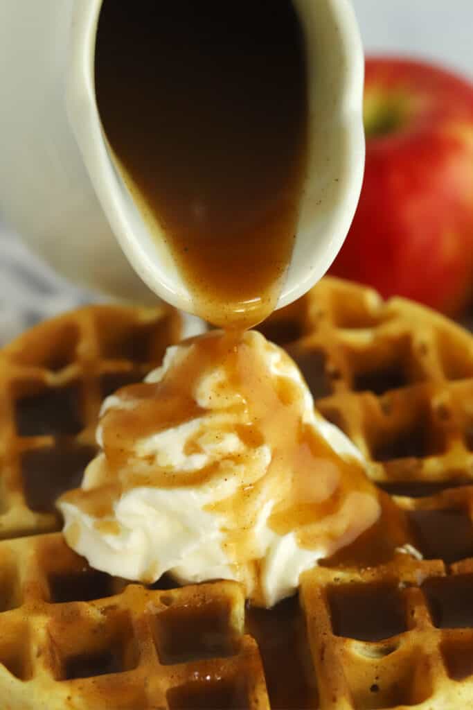 Apple brown sugar syrup poured from a carafe over waffles and whipped cream.
