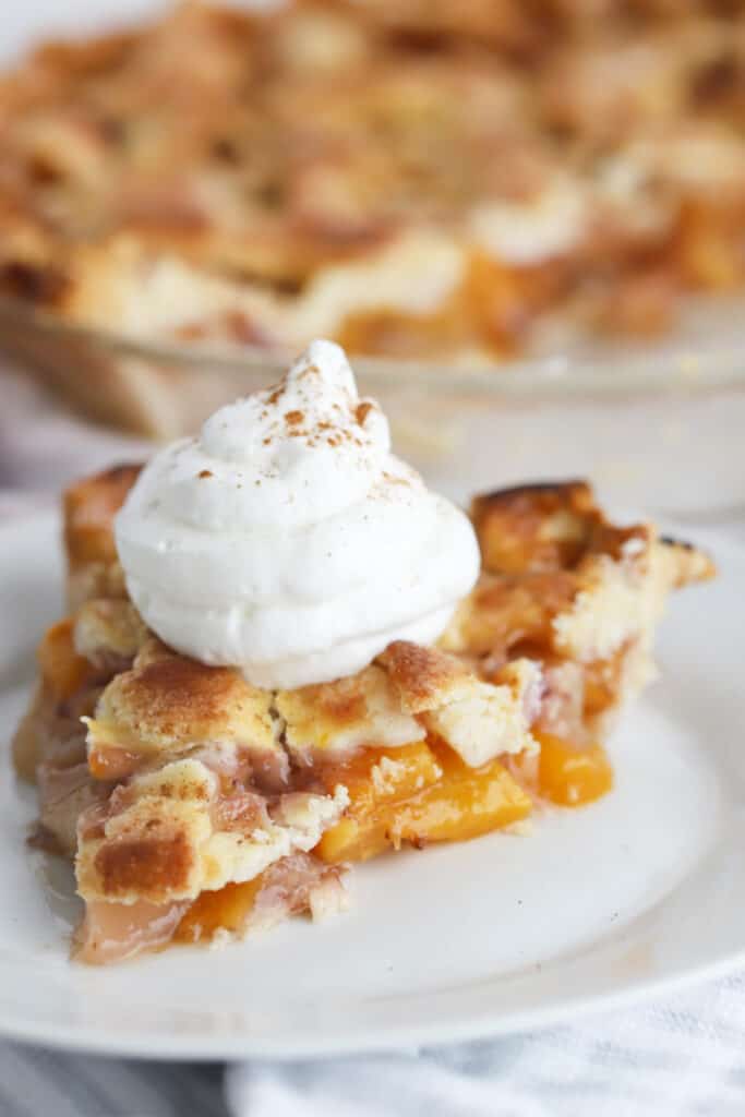 A slice of peach pie on a white plate topped with fresh whipped cream.