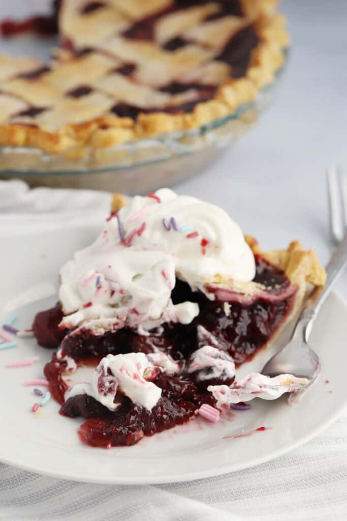 A slice of cherry pie topped with whipped cream on a white plate with a fork.