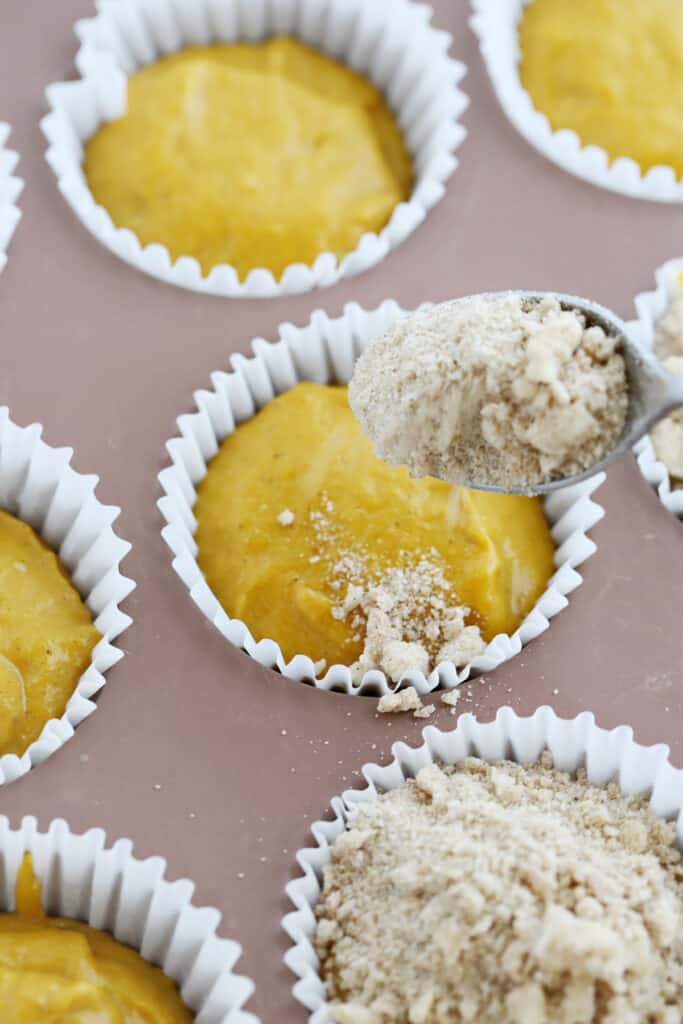 Muffin tins full of batter with streusel being spooned over the top, easy pumpkin muffin recipe. Spice cake pumpkin muffins.