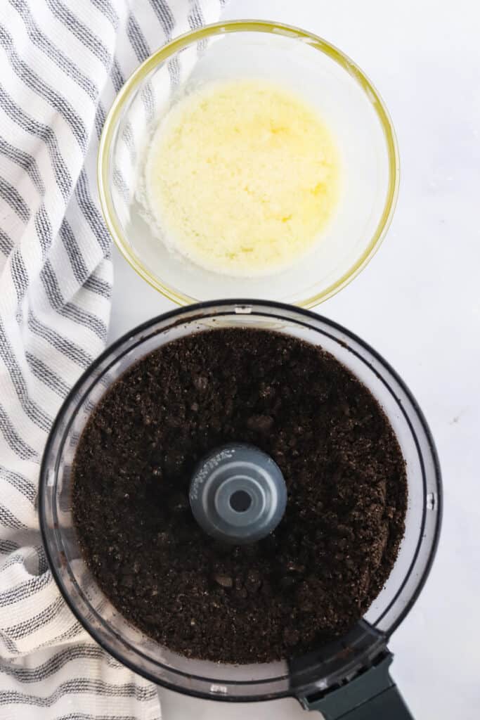 A food processor full of Oreo crumbs and a small bowl full of melted butter on a table.
