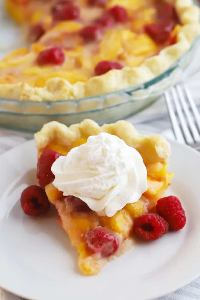 fresh peach and raspberry pie recipe on a white plate with fresh whipped cream. a favorite summer pie recipe.