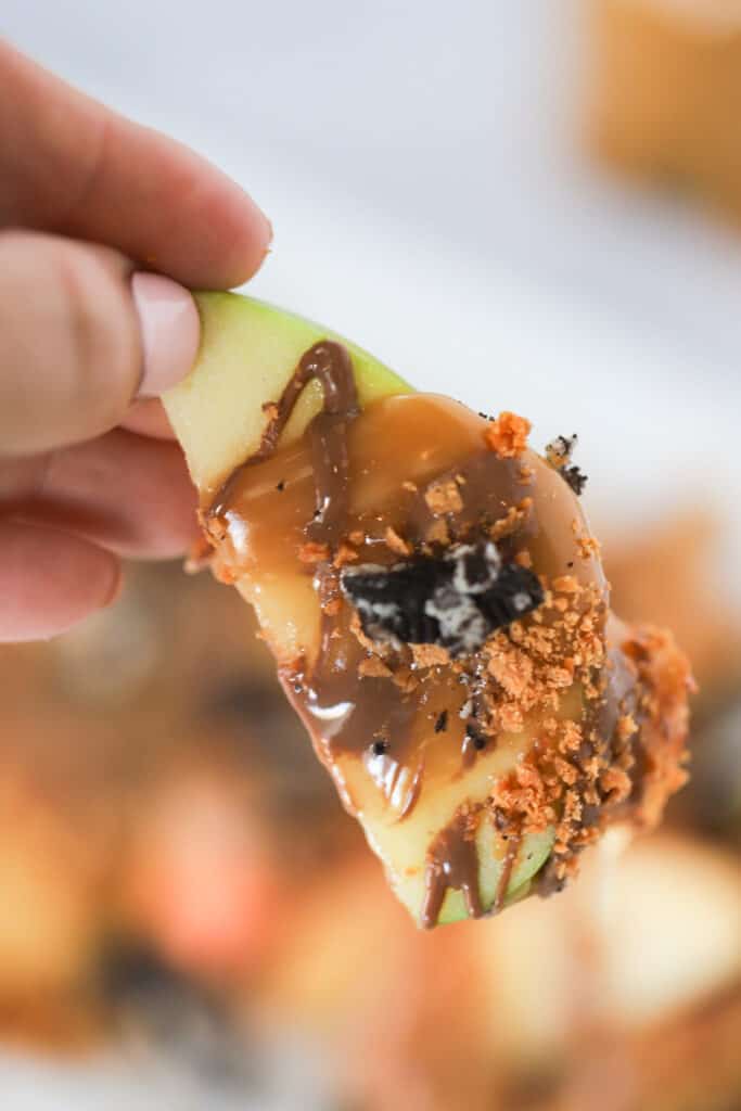 A hand holding an apple slice covered in caramel sauce and chopped candy.