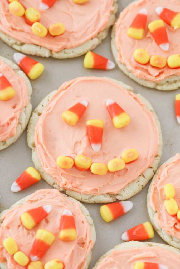 Baked cookies on a baking sheet topped with orange frosting and decorated with candy corns to look like jack-o'-lanterns.