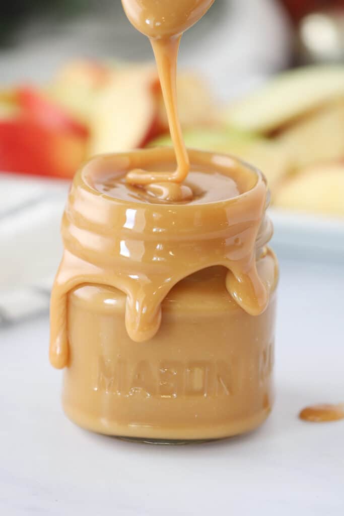 A small mason jar overflowing with gooey caramel dip recipe for apples.
