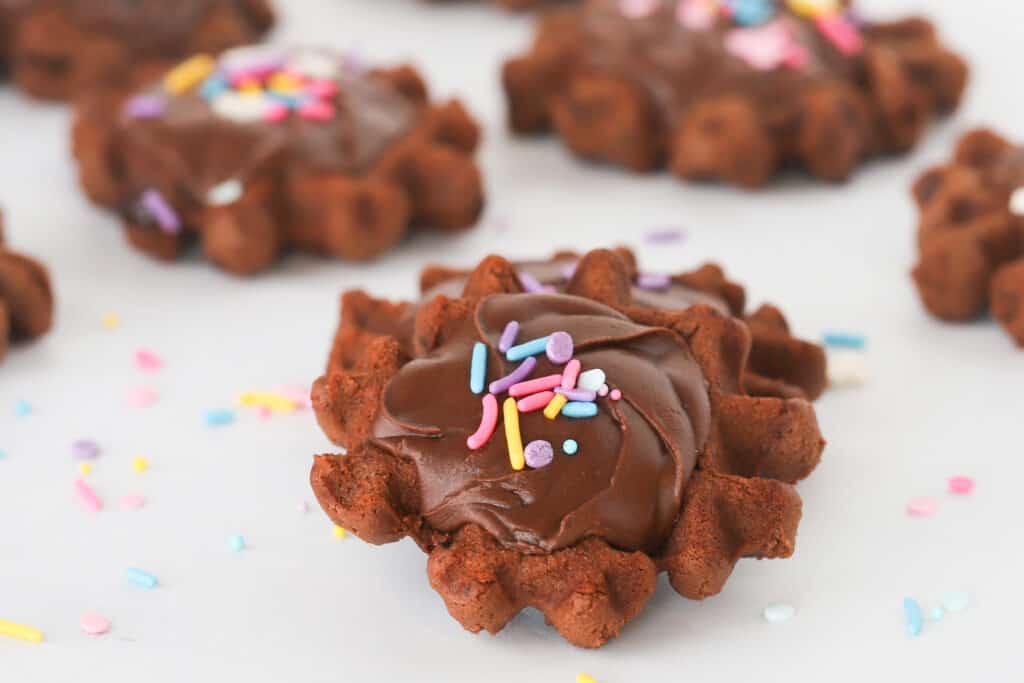 Waffle brownie cookies on a table topped with chocolate frosting and sprinkles.