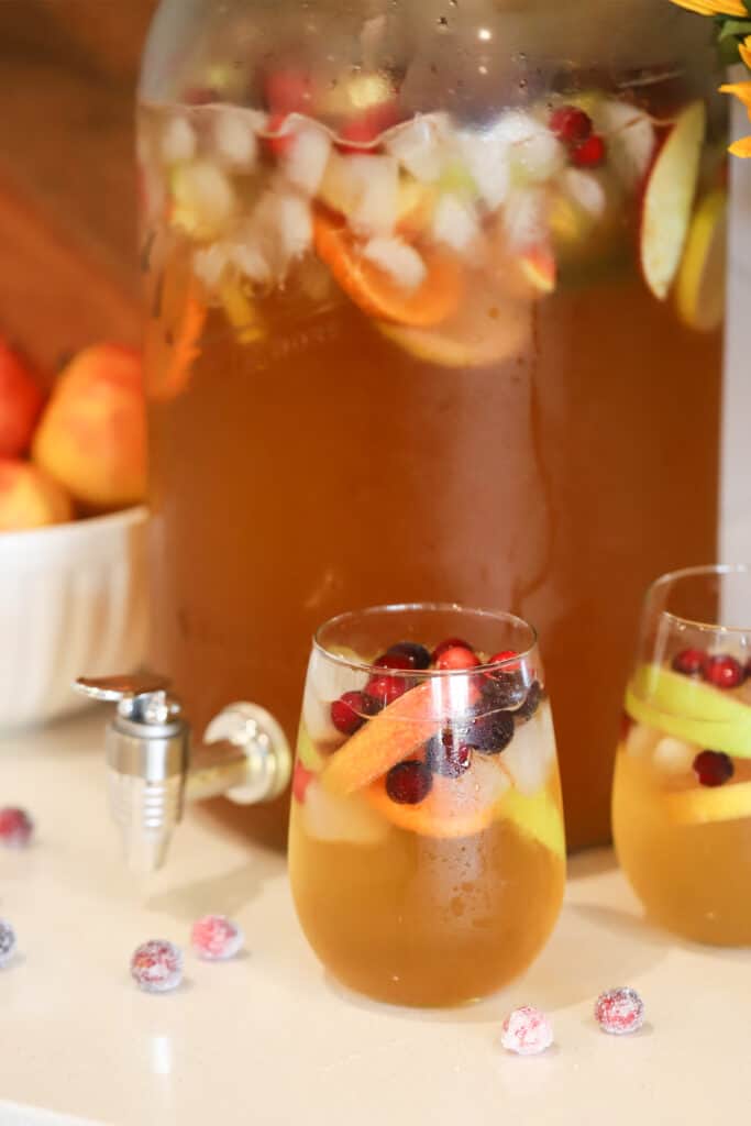 A large glass pitcher and two individual glasses full of apple punch that has been garnished with apple slices and cranberries.