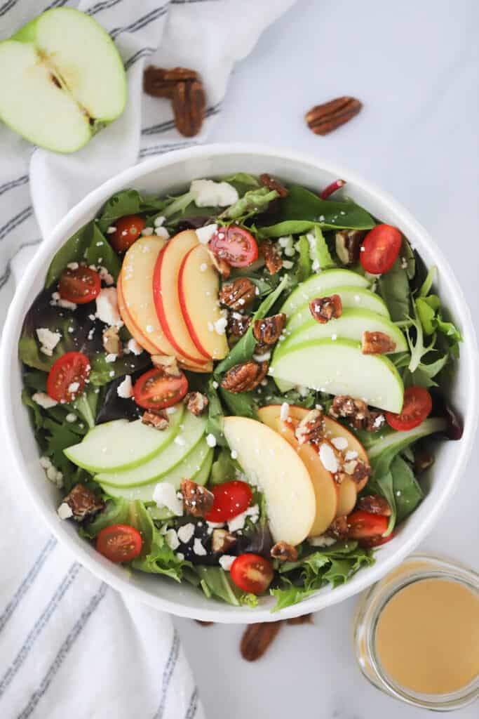 A bowl full of salad with greens, apples, tomatoes and pecans.