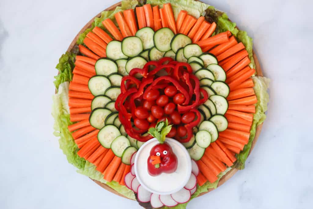 A thanksgiving veggie platter made with carrots, cucumbers, bell peppers, radishes and cherry tomatoes with a ramekin of dip decorated with a veggie face.