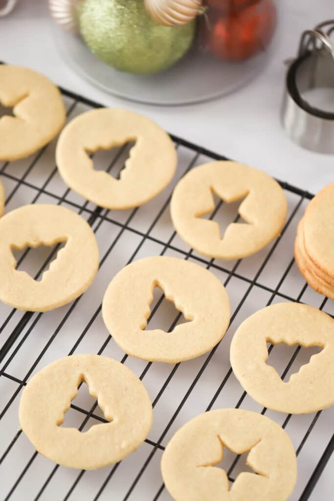 Round cookies with tiny Christmas trees and stars cut out of the centers, laying on a wire cooling rack.