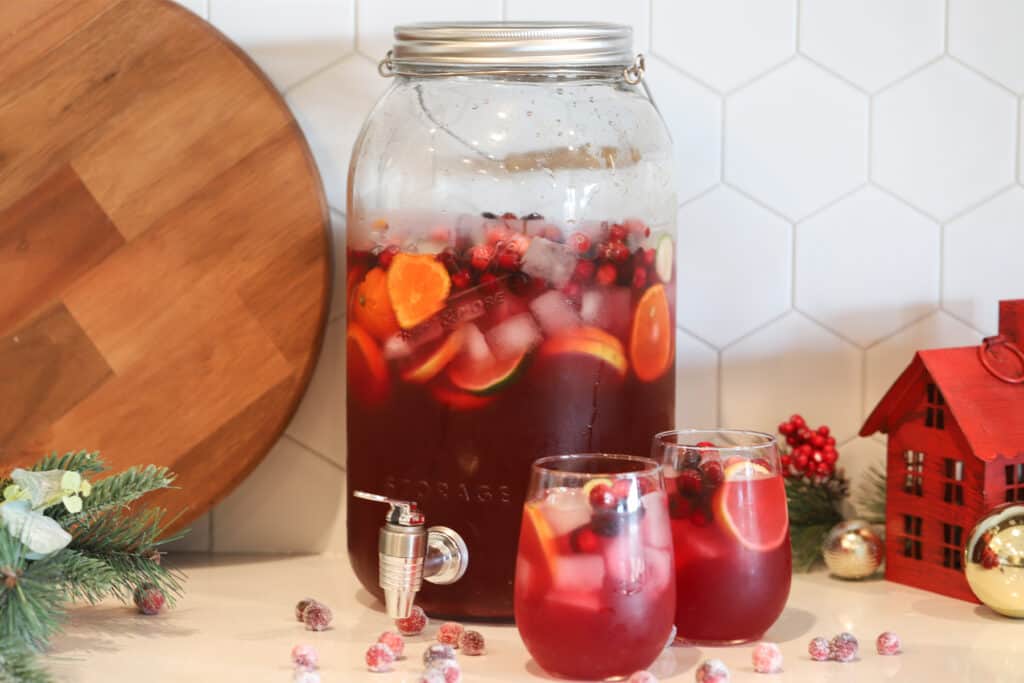 A table with a large drink dispenser and two individual glasses full of red punch, garnished with sliced fruit and cranberries. Cranberry ginger ale punch. Ginger ale punch recipe. Punch with ginger ale.