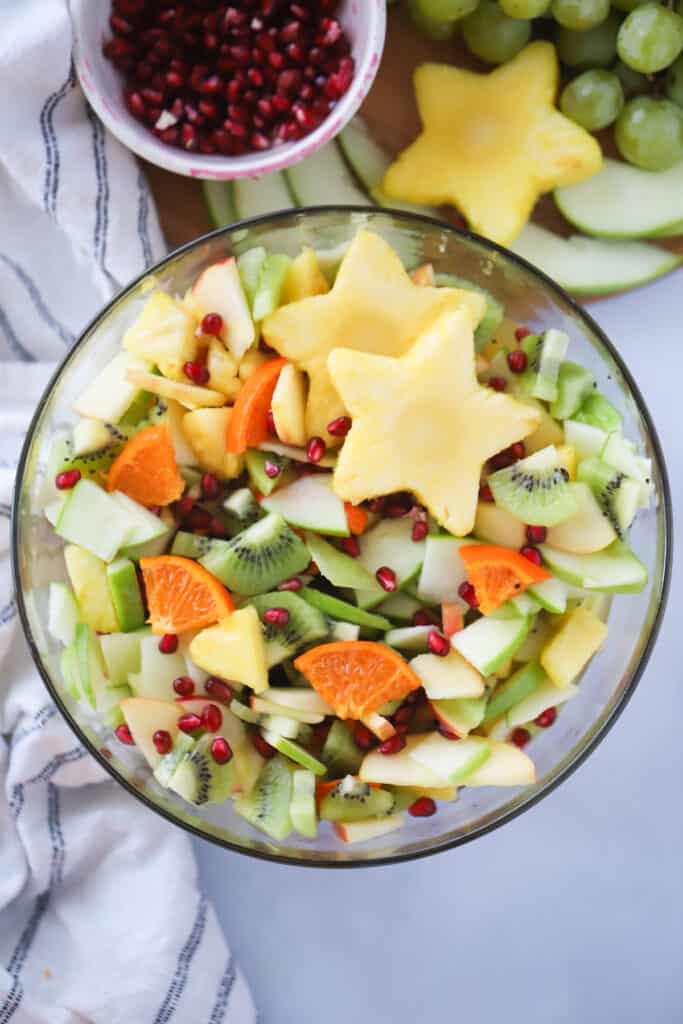 A large glass serving bowl full of winter fruit salads on a table next to a small bowl full of pomegranate seeds. Fruit salad recipe winter. Fall fruit salad. holiday fruit salad. kiwi fruit salad. pomegranate fruit salad. fruit salad with pomegranate.