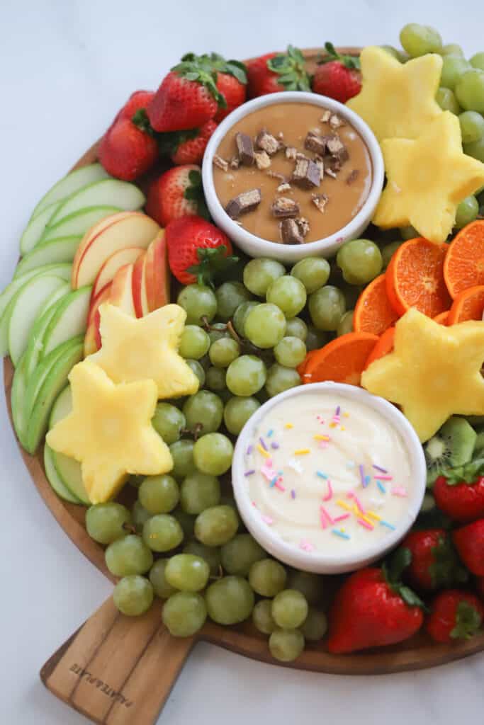 Fresh fruit served on a round wooden board with small ramekins of dips for serving.