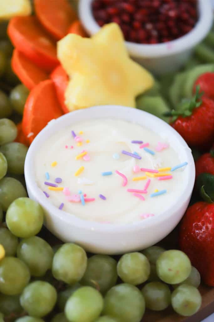A small white ramekin with cream cheese dip garnished with sprinkles, on top of a plate full of fresh fruit.