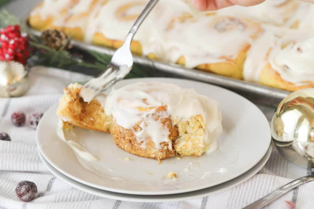 A cinnamon roll on a white plate being eaten with a fork, with a full tray of cinnamon rolls in the background.