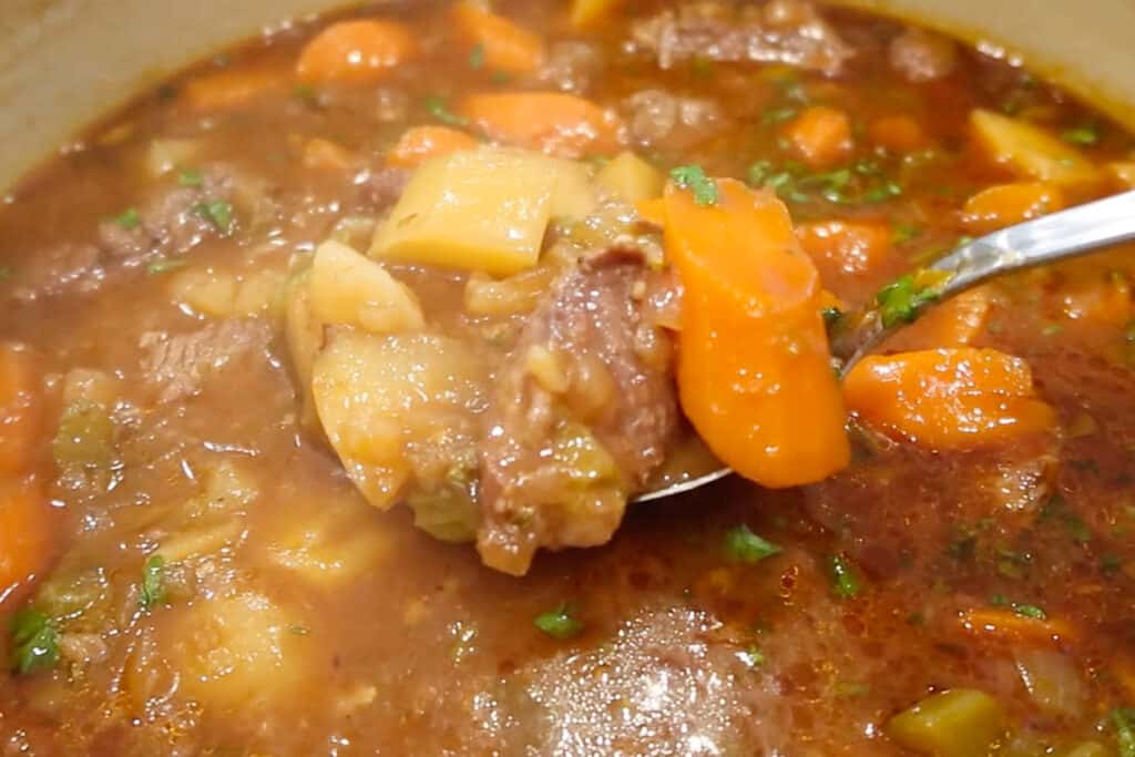 A bowl full of beef stew recipe and a spoon holding chunks of cooked carrots, potatoes and beef_stew.