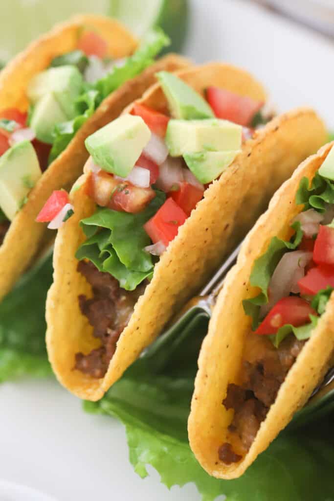Hard taco shells resting in a taco holder, filled with taco meat, shredded lettuce, diced tomatoes and more, best taco recipes round beef